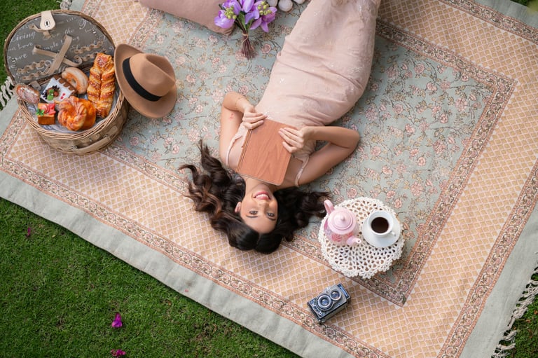 Woman lying on blanket outdoors with picnic setup, surrounded by basket of food, drinks, and casual items on grass