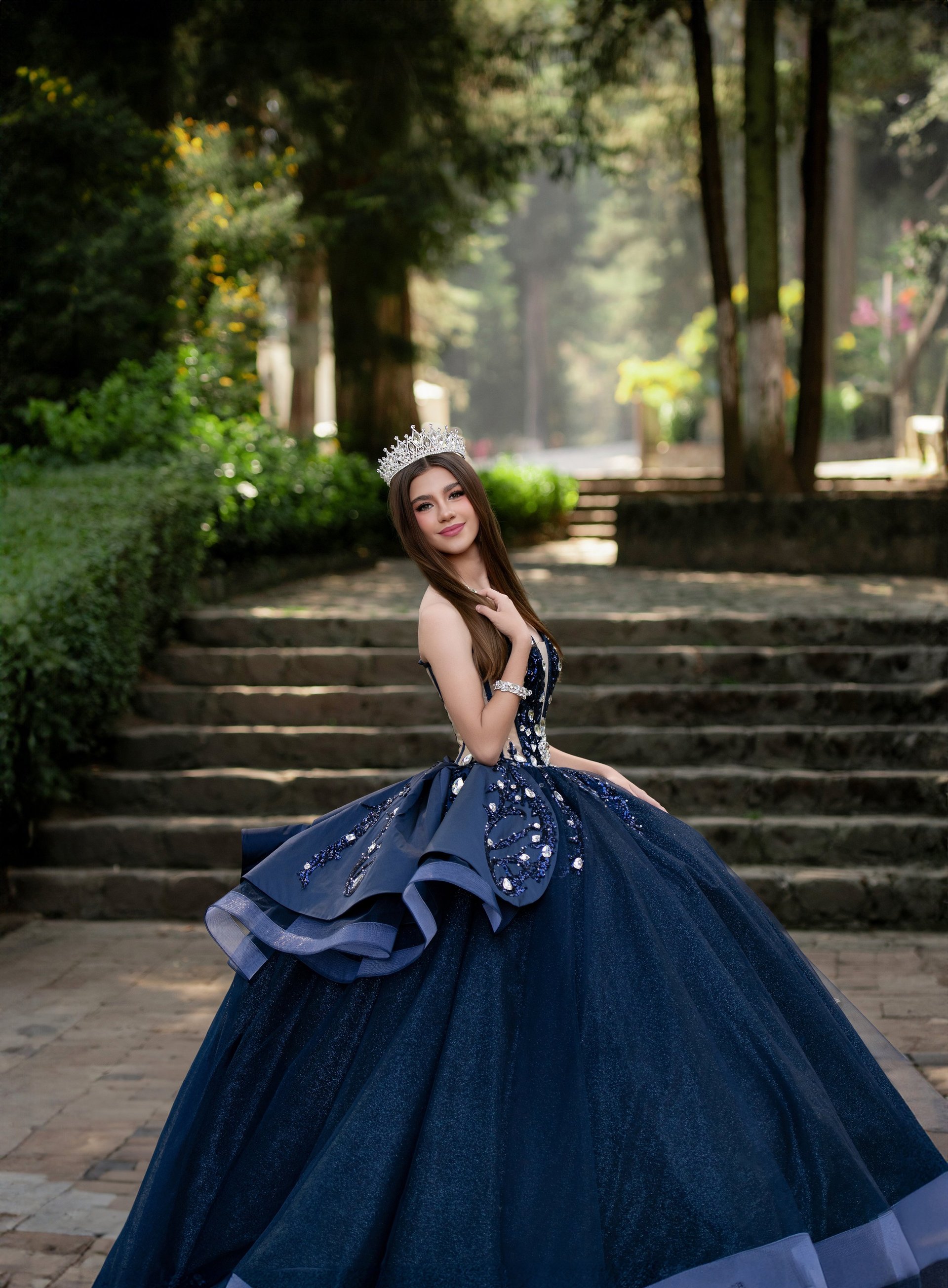Young woman in navy ball gown and tiara posing on tree-lined pathway with stone steps