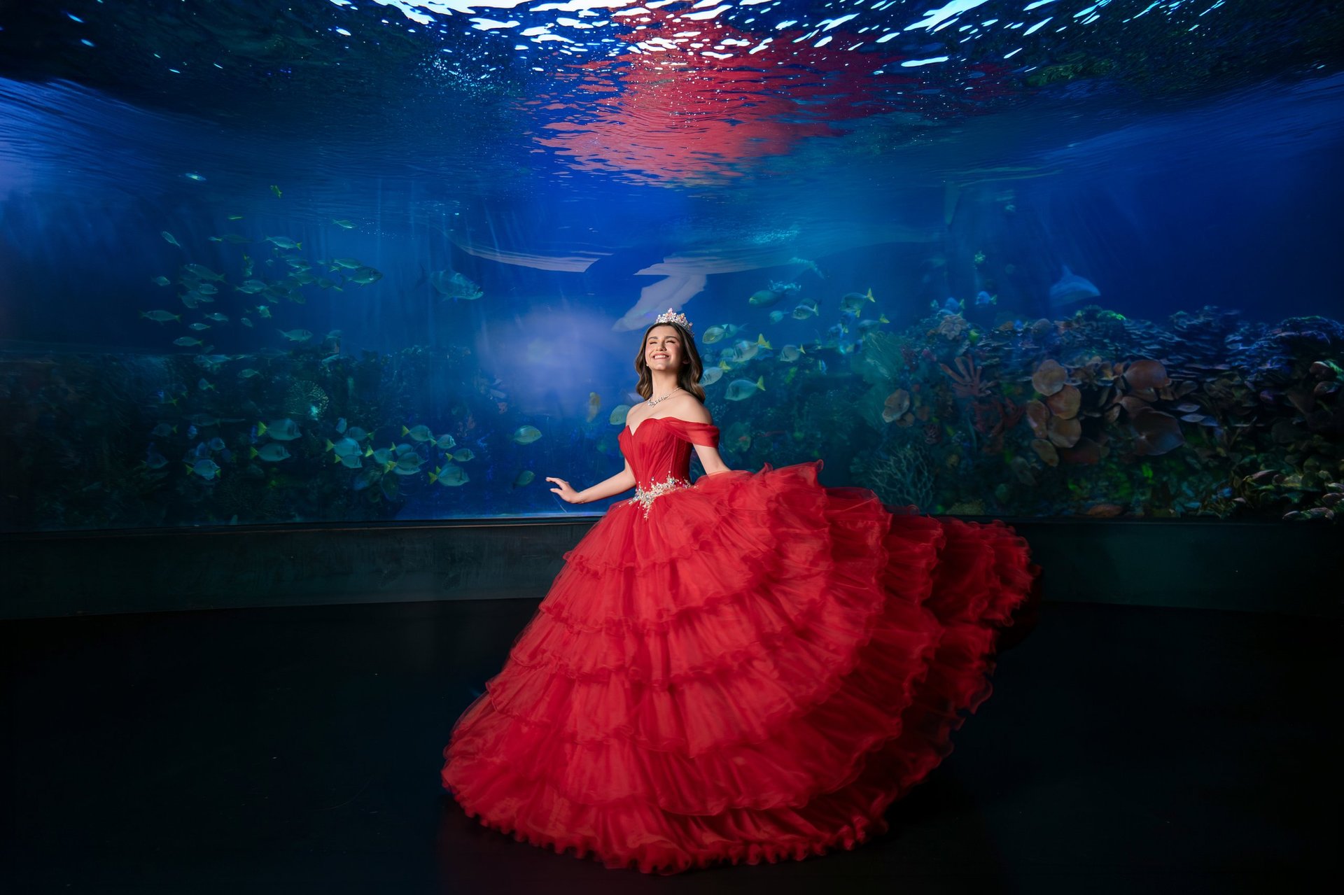 Woman in flowing red ball gown stands underwater in an aquarium surrounded by marine life and coral