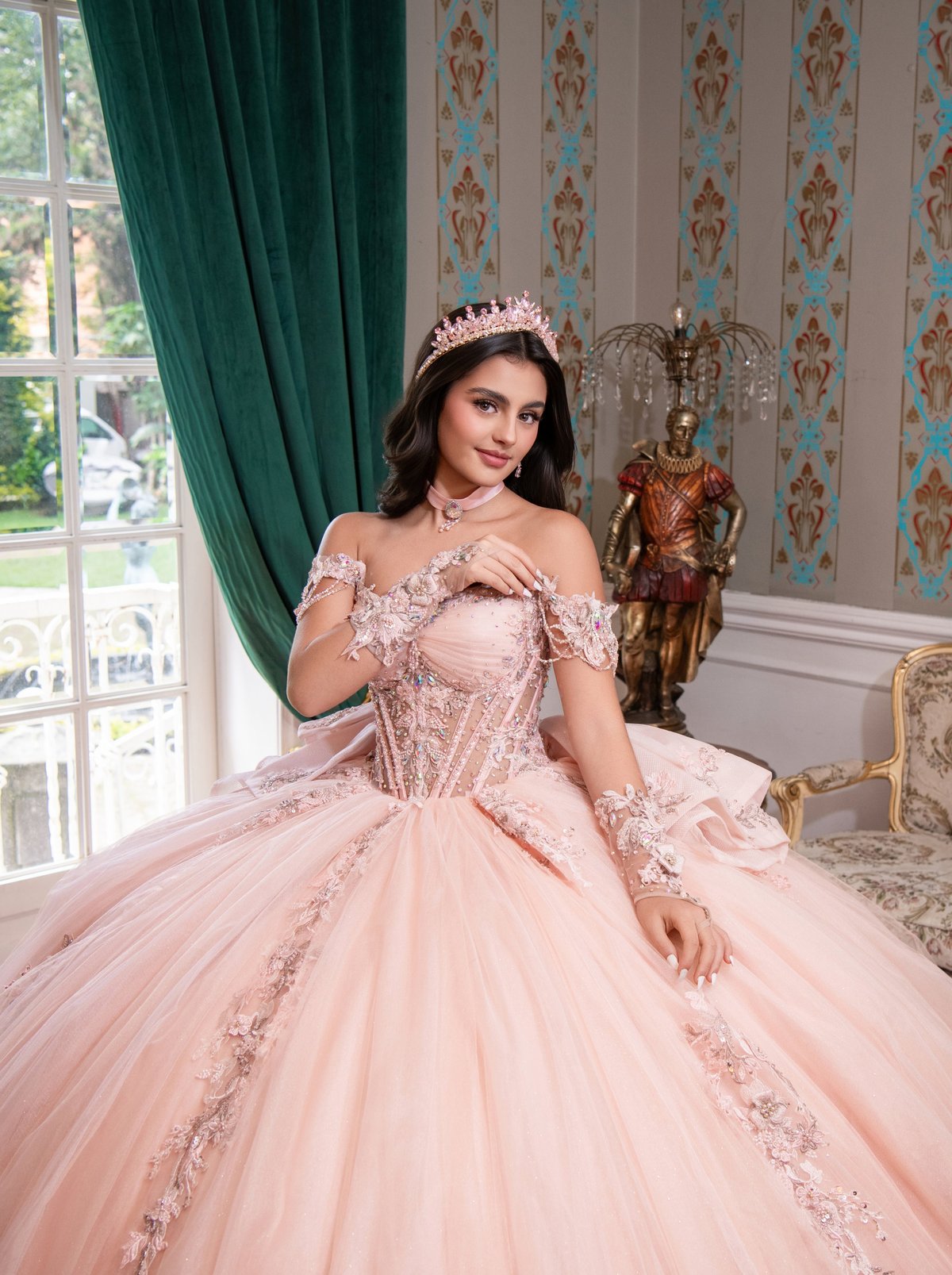 Young woman in an ornate pink ball gown with tiara and off-shoulder design, posed in an elegant palace setting with green curtains and patterned wallpaper