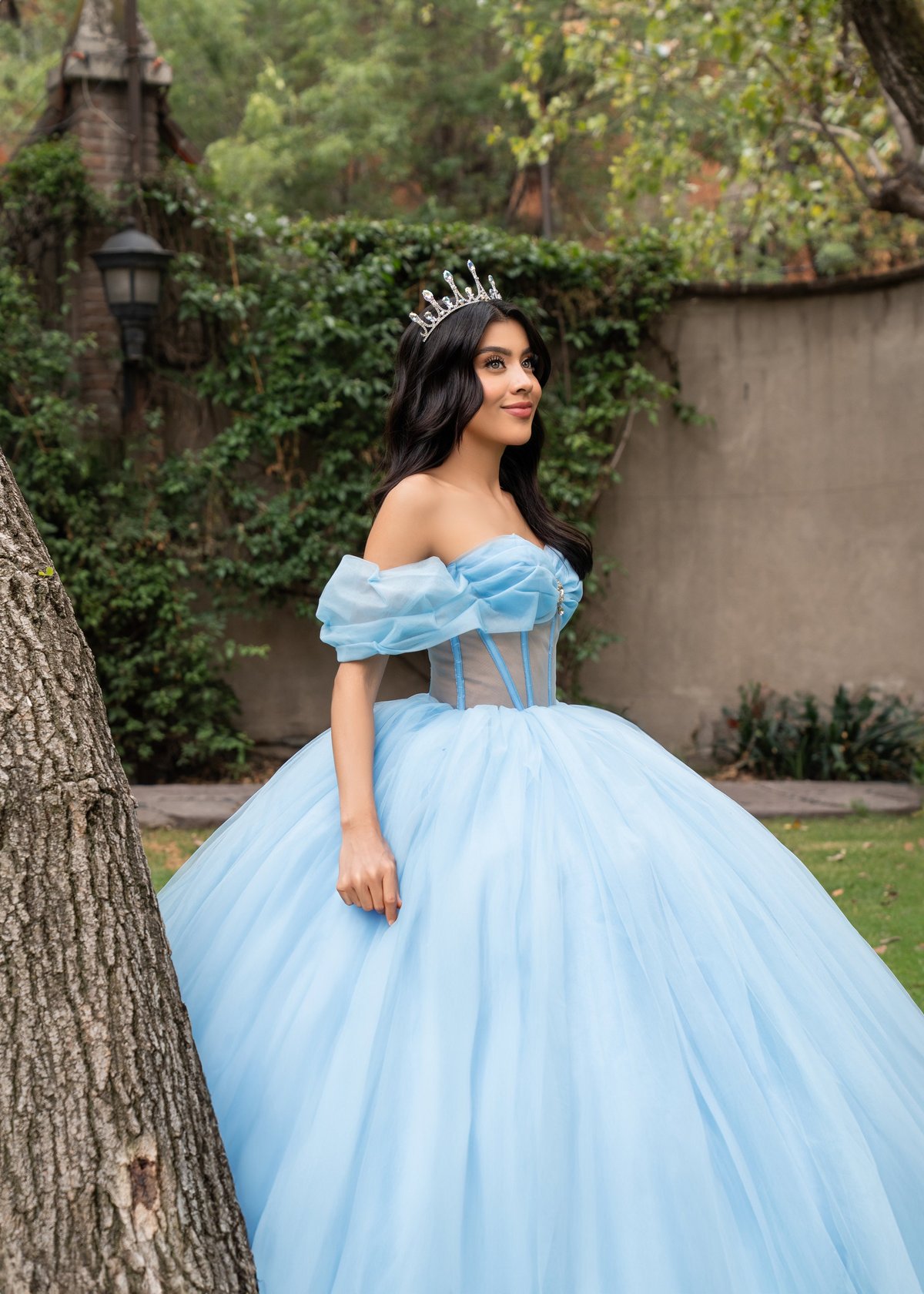Woman in light blue off-shoulder ball gown with tiara standing against ivy-covered wall and tree trunk