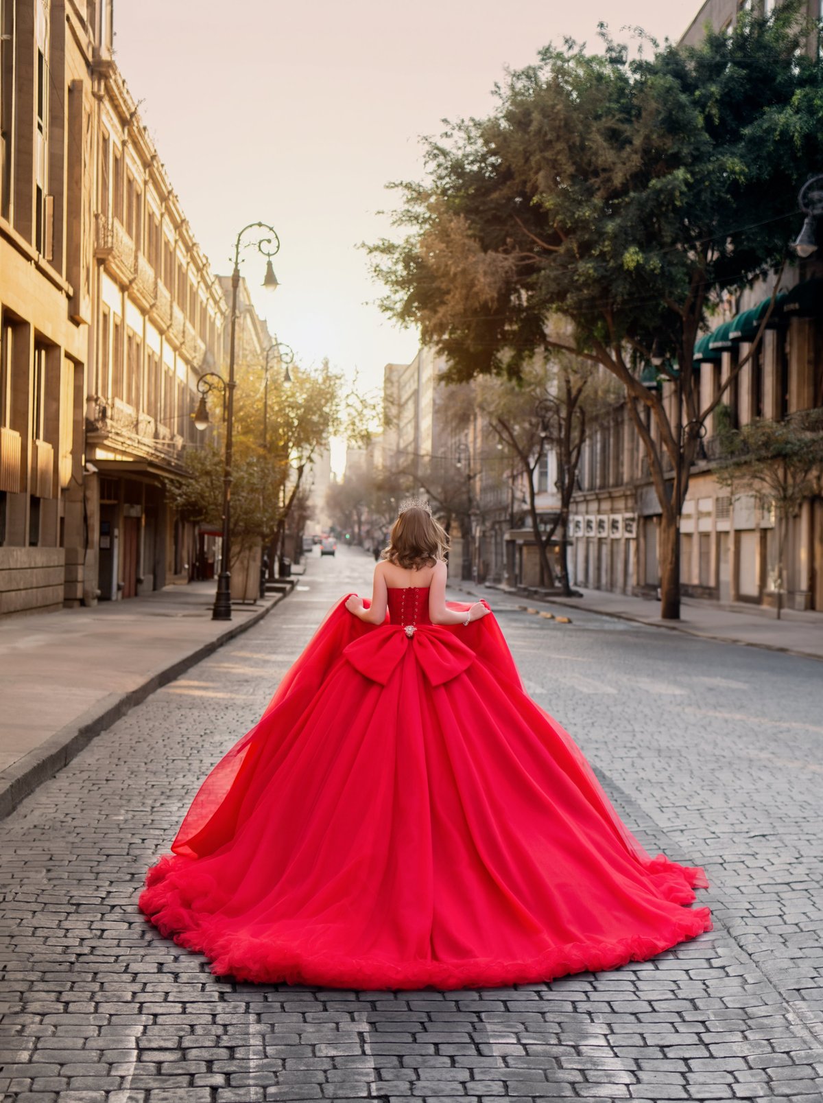 Woman in red ball gown walking down tree-lined European street at sunset