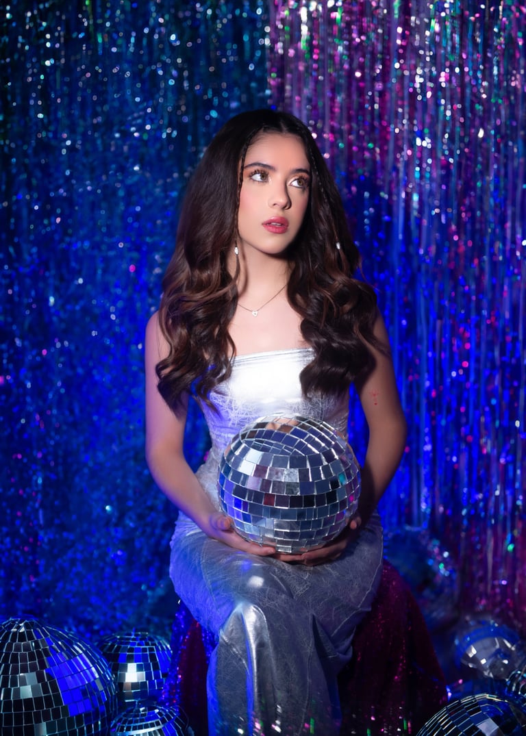 Woman in white dress holding a silver disco ball against a blue and purple sequined backdrop with matching disco balls at her feet